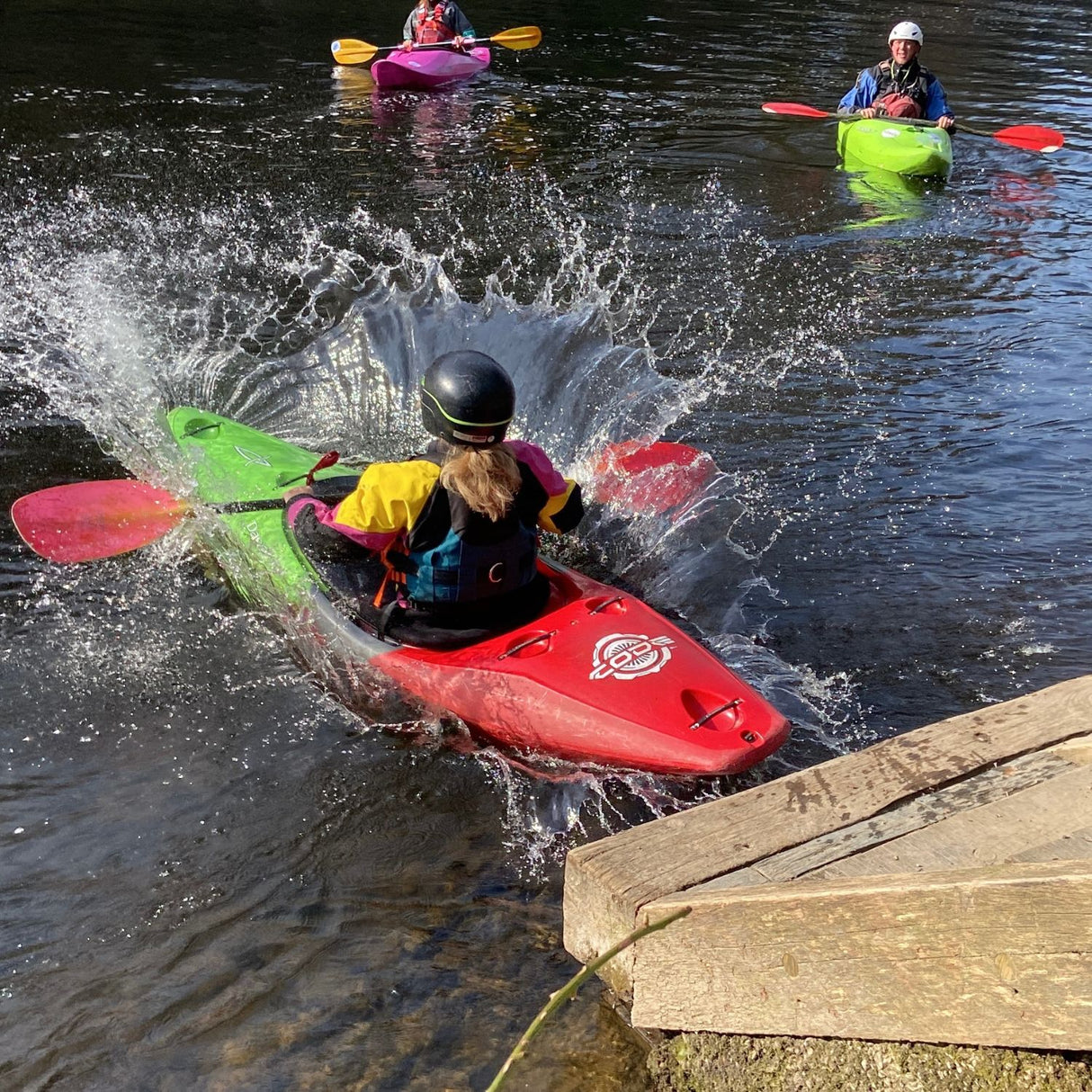 Person kayaking in a red kayak with water splashing around, near a wooden dock.