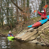 Two people in kayaks at the edge of a wooden platform with trees in the background