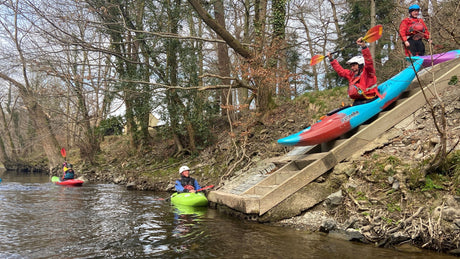 People preparing to kayak by a river with trees and a stone staircase in the background.